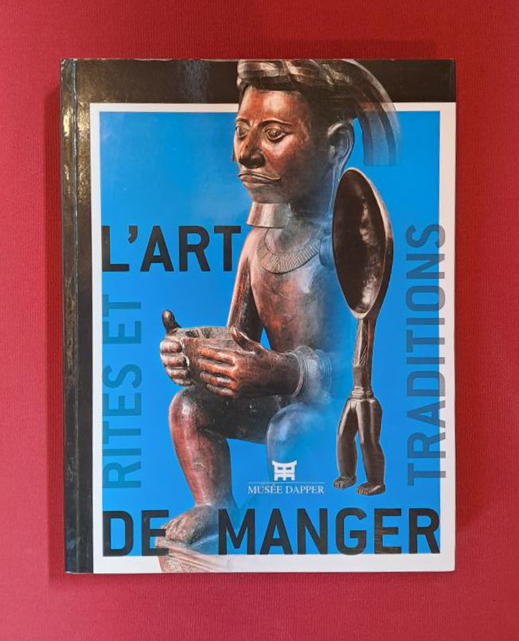 L'Art de Manger. The book cover is bright blue, with a wooden carved statue of a human and another carving in the centre. The book is set on a dark red background