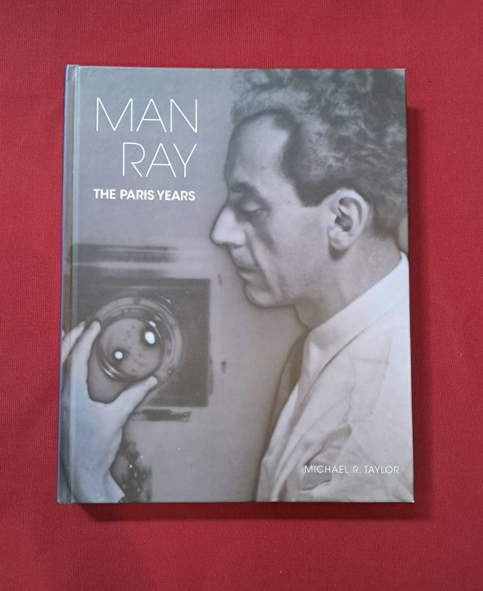 Man Ray. Cover of book shows black and white image of a man adjusting a camera from side perspective. Set on a dark red background