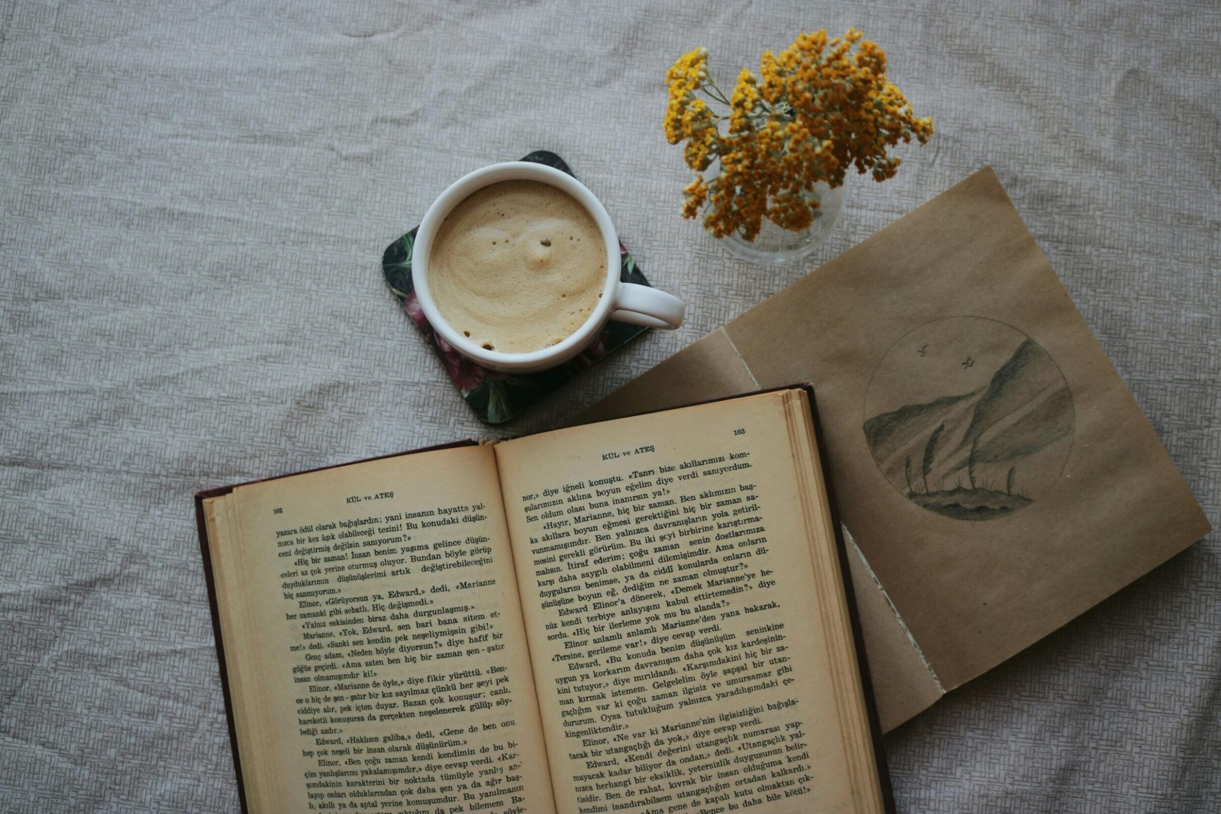 A book lies open, with tanned brown pages. Next to it is a black and white drawing of mountains. In the upper part of the image is a vase of yellow flowers, and a mug of foamy coffee